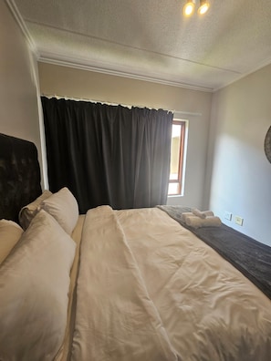 A hotel room with two beds, a window with curtains, and a wall-mounted light fixture. - Cozy One-Bedroom Apartment In Fourways (Sandton)