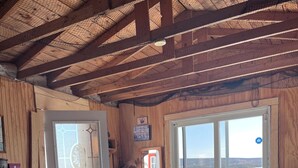 Interior de una cabaña de madera con una cama, un aparador y vistas del exterior a través de una puerta.