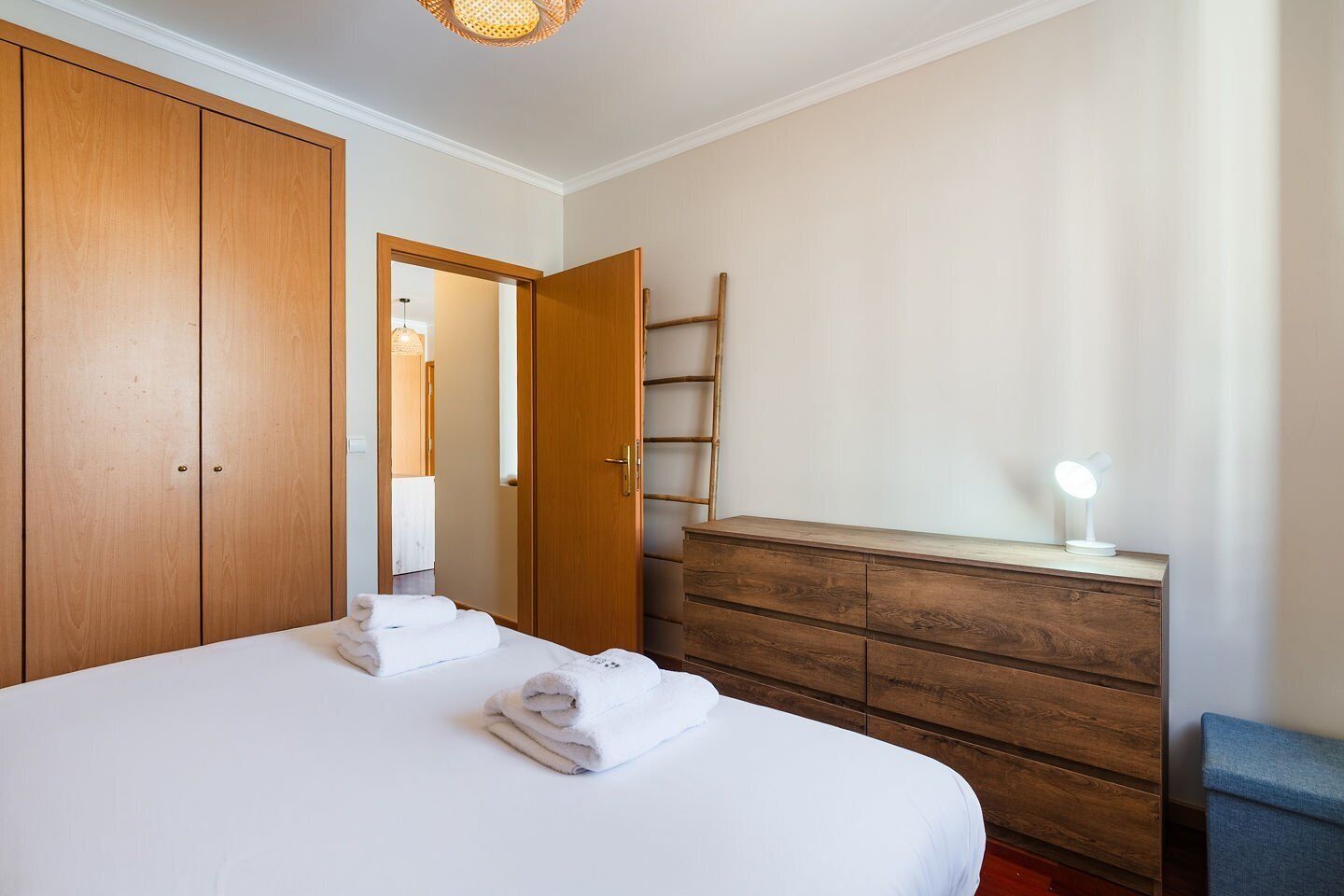 A bedroom with a bed, wooden wardrobe, dresser, and a bedside lamp.