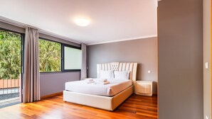 A modern bedroom with a large bed, a nightstand, a ceiling light, and a view of greenery outside.