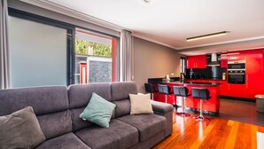 A modern living room with a grey sofa, a kitchen with red cabinets, and a large window.