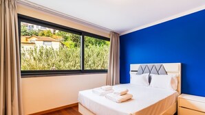 A hotel room with a bed, a nightstand, a window with curtains, and a view of greenery outside.