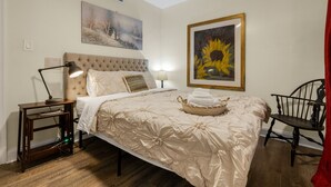 A bedroom with a tufted headboard, a bed with a beige comforter, a bedside lamp, a framed painting of a sunflower, and a wooden chair. - Garden Oasis Room near Wynwood (Miami)