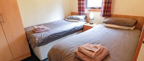 Ein Zimmer mit zwei Betten, einem Holzschrank, einem Nachttisch mit Lampe und einem Fenster mit Vorhängen.