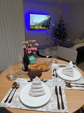 A dining table set with plates, cutlery, a cake, a vase with flowers, and a television in the background.