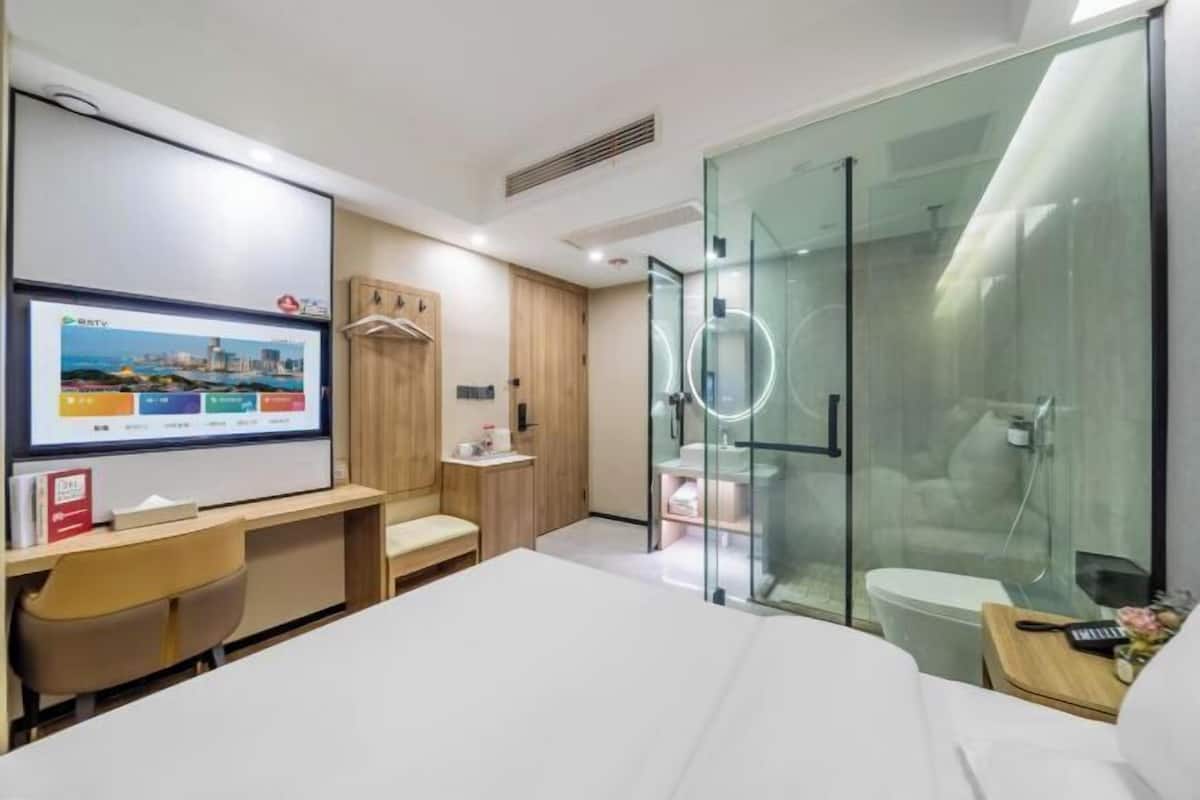 Una habitación de hotel moderna con una cama grande, un escritorio con una computadora, cabina de ducha de vidrio y un televisor que muestra un paisaje urbano.