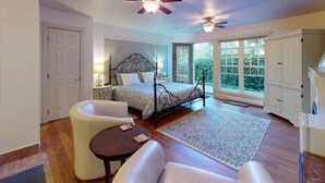 A bedroom with a bed, a chair, a table, a ceiling fan, and a view of greenery outside. - Annapolis Suite - Private HotTub, KingBd, DT (Wolfville)