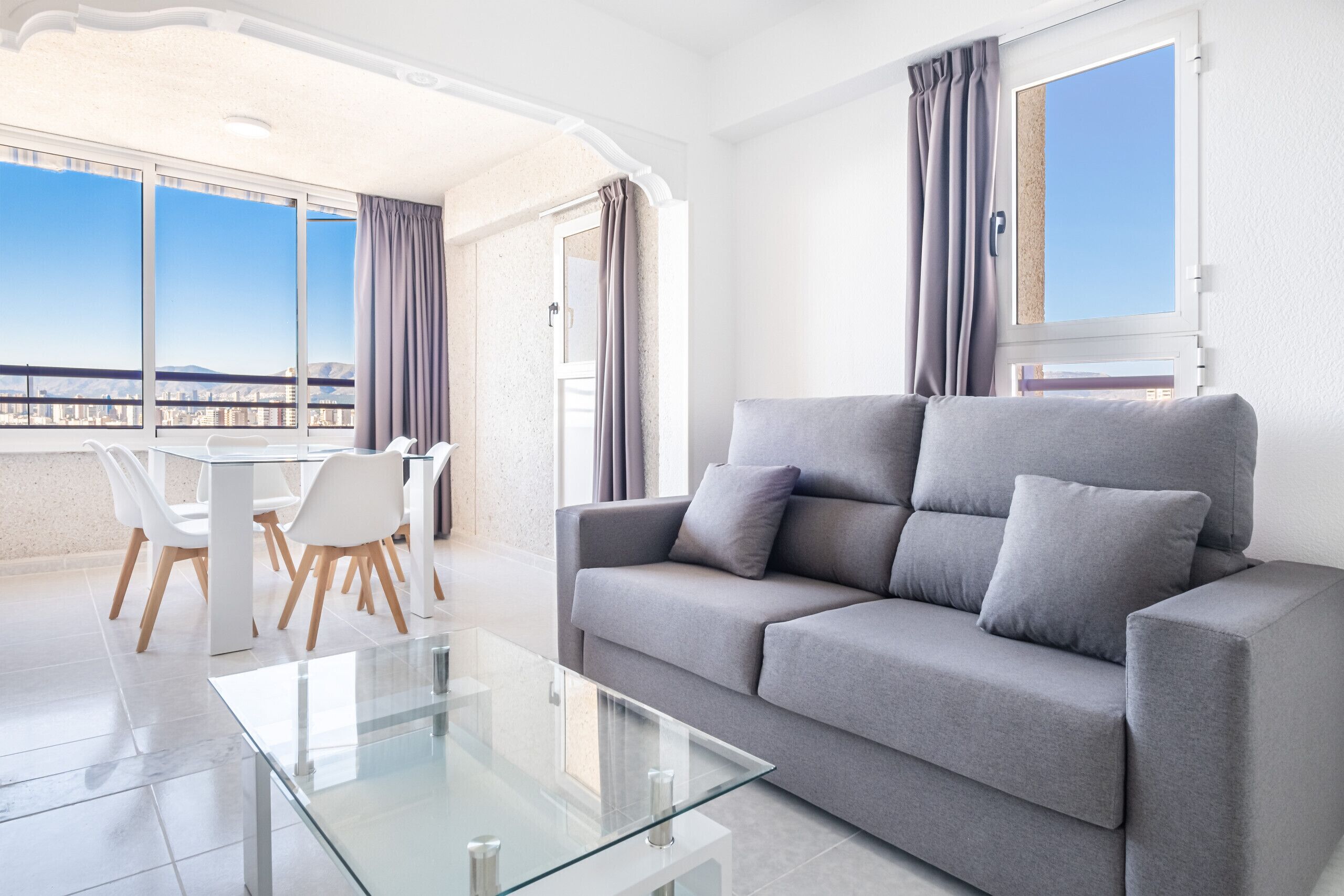 A modern living room with a grey sofa, a glass coffee table, and a dining area with white chairs.