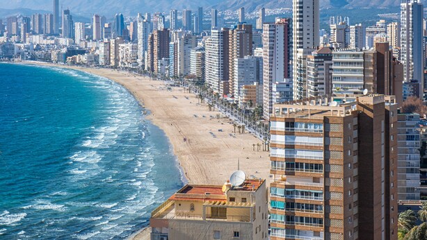 Paisagem costeira com edifícios altos, uma praia e montanhas ao fundo.