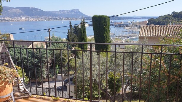 En balkon med udsigt over en lystbådehavn, et bjerg i baggrunden og en bygning med tegltag.