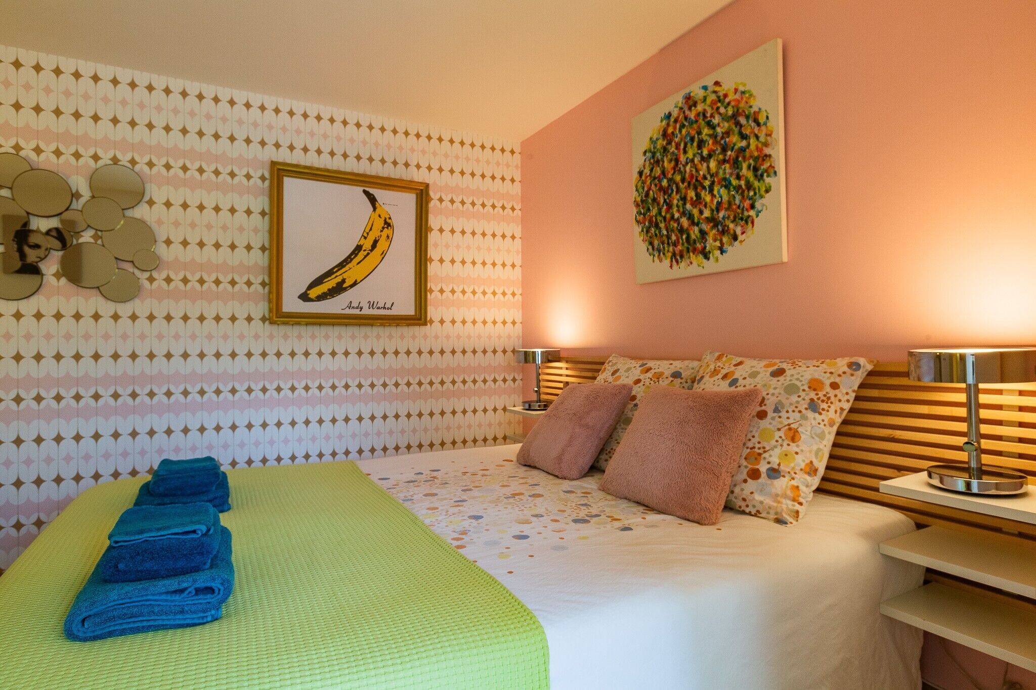 A neatly made bed with a green bedspread, a framed banana artwork, a colorful abstract painting, and a wall with a geometric pattern.