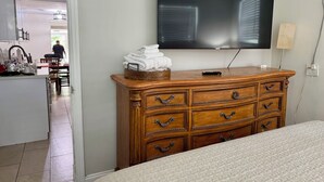 Une chambre à coucher avec un lit, une commode en bois dotée de tiroirs, un téléviseur fixé au mur et un coin cuisine visible à travers une porte ouverte.