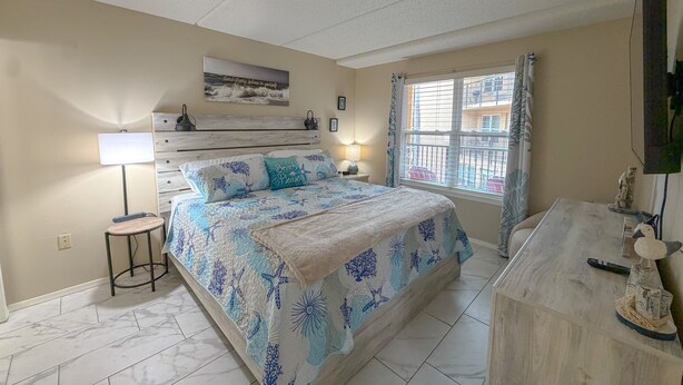 A bedroom with a bed, bedside lamp, and a window with a view of buildings. - Pool- BeachSide- Pet Friendly- 2-3min walk to beach. (South Padre Island)