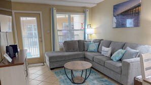 A living room with a grey sectional sofa, a round coffee table, a blue patterned rug, and a painting of a pier on the wall. - Pool- BeachSide- Pet Friendly- 2-3min walk to beach. (South Padre Island)