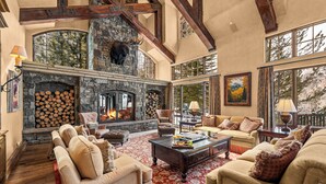 A spacious living room with a stone fireplace, wooden beams, and large windows.