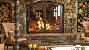 A stone fireplace with logs, leather armchairs, a coffee table with fruit and drinks, and a view into the room.