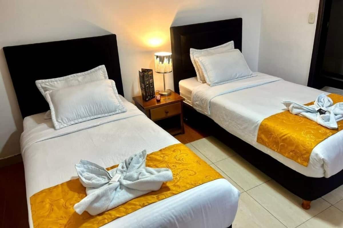 Dos camas de hotel contiguas con sábanas blancas, una mesita de noche con lámpara y una toalla sobre la cama.