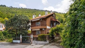 Una casa de dos pisos con un balcón, rodeada de abundante vegetación y colinas.