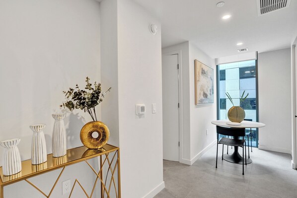 Un couloir avec un mur blanc, une console dorée ornée de vases, une table ronde avec un vase, et une porte donnant sur une autre pièce.