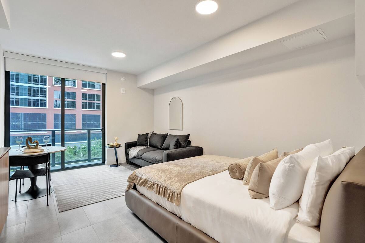Quarto de hotel moderno com uma cama grande, um sofá, uma mesa de jantar e vista para um edifício por uma grande janela.