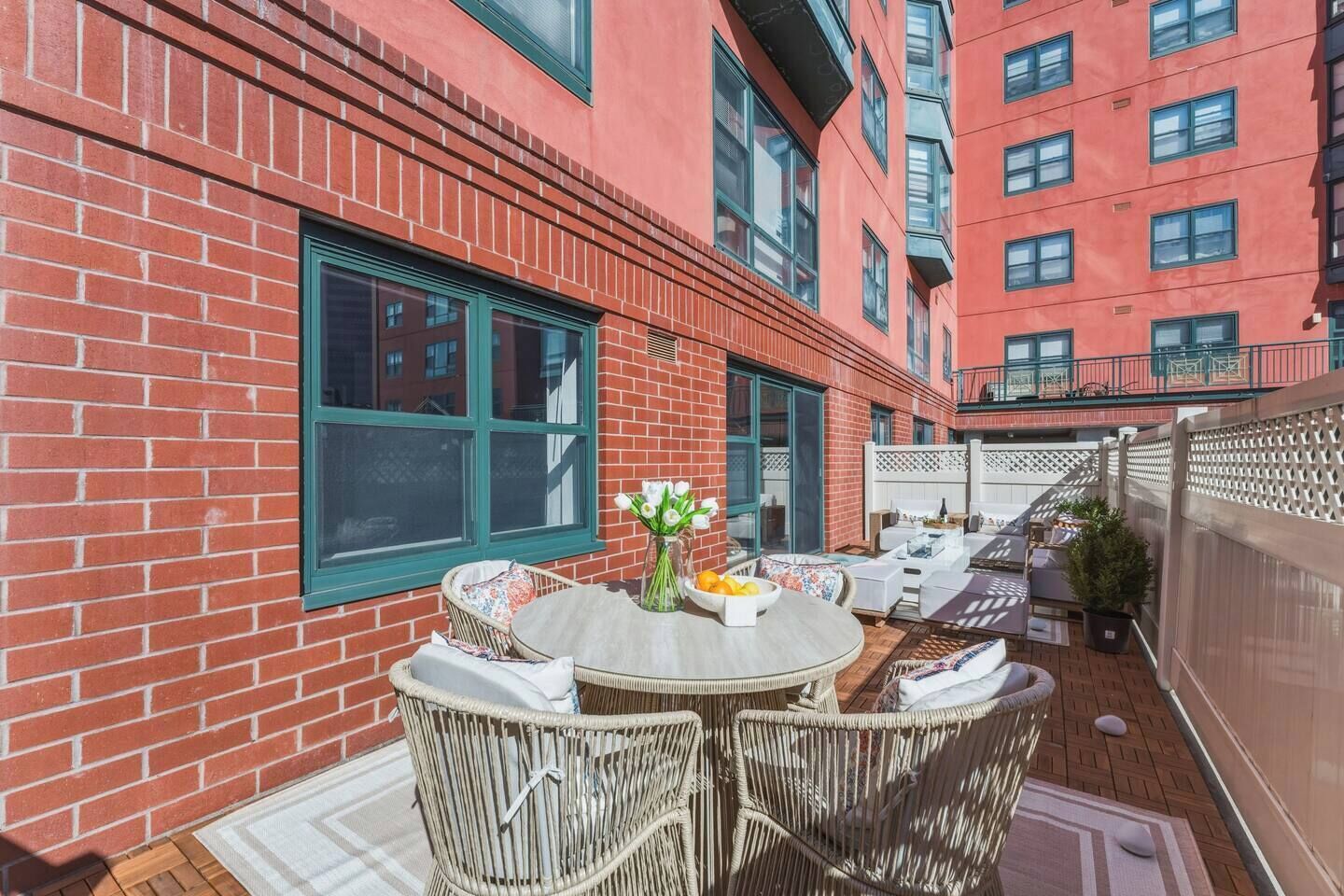 A patio with wicker furniture, a table with a vase of flowers, and a brick building with large windows.