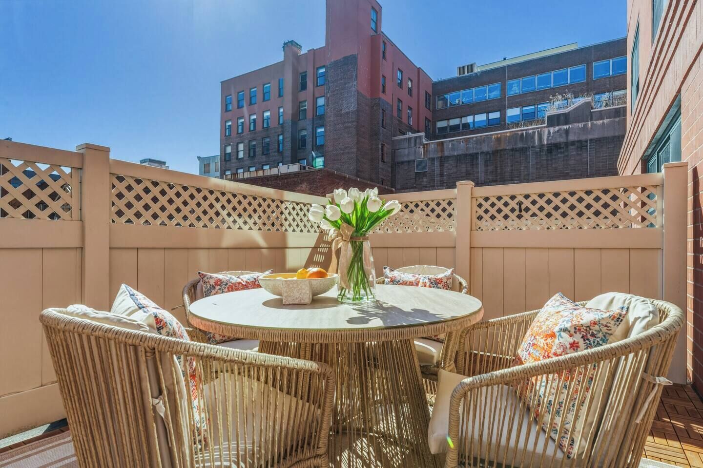 A round table with wicker chairs, a vase with flowers, and a bowl of fruit on a balcony with a view of buildings.