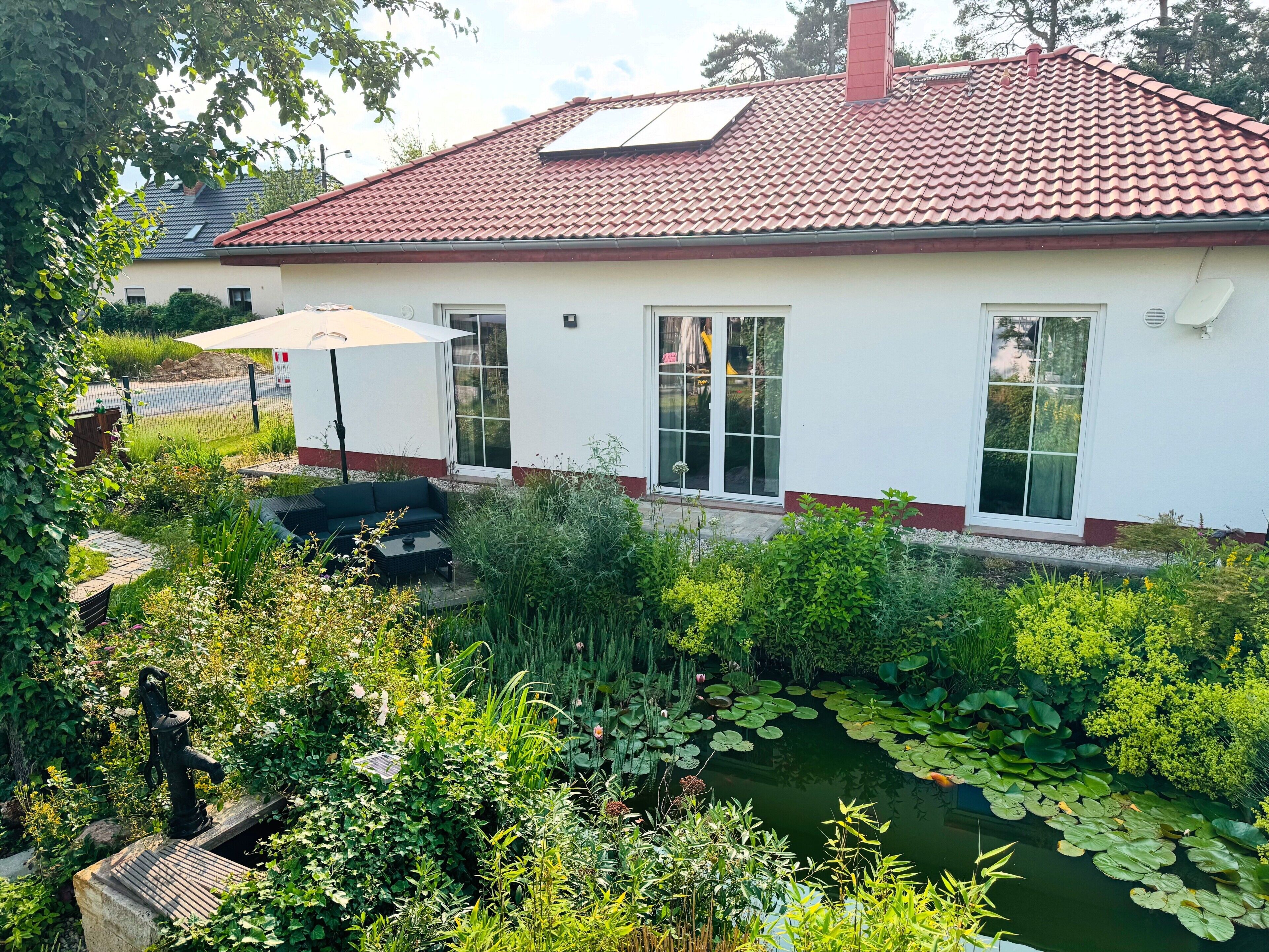 Ein Haus mit rotem Ziegeldach, weißer Fassade und einem Garten mit Teich und Grünpflanzen.
