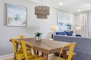 A dining area with a wooden table and yellow chairs, a rattan chandelier, and a grey sofa with blue cushions. - Oceanfront NEW Updated Modern Condo w/ 4 Beds! (North Myrtle Beach)