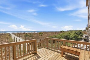 Een houten terras met een bankje, met uitzicht op een strand met een houten omheining en begroeiing.