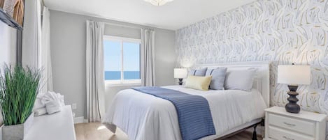 Ein Schlafzimmer mit einem Bett, Nachttischen, einer Lampe und einem Fenster mit Vorhängen, mit Blick auf das Meer.