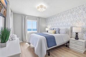 A bedroom with a bed, bedside tables, a nightstand, a lamp, a window with curtains, and a view of the sea. - Oceanfront NEW Updated Modern Condo w/ 4 Beds! (North Myrtle Beach)