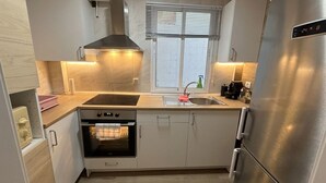 Ett kompakt kök med en inbyggd ugn, ett kylskåp i rostfritt stål och ett fönster med persienner.