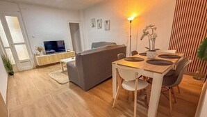 Ett modernt vardagsrum med en soffa, ett matbord och stolar, en tv placerad på ett träskåp och en krukväxt.