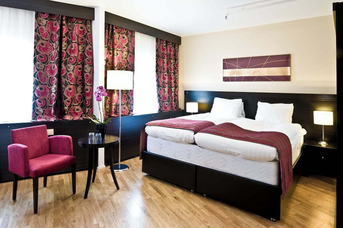A hotel room with a bed, two bedside tables, a chair, a small table with a lamp, and curtains.