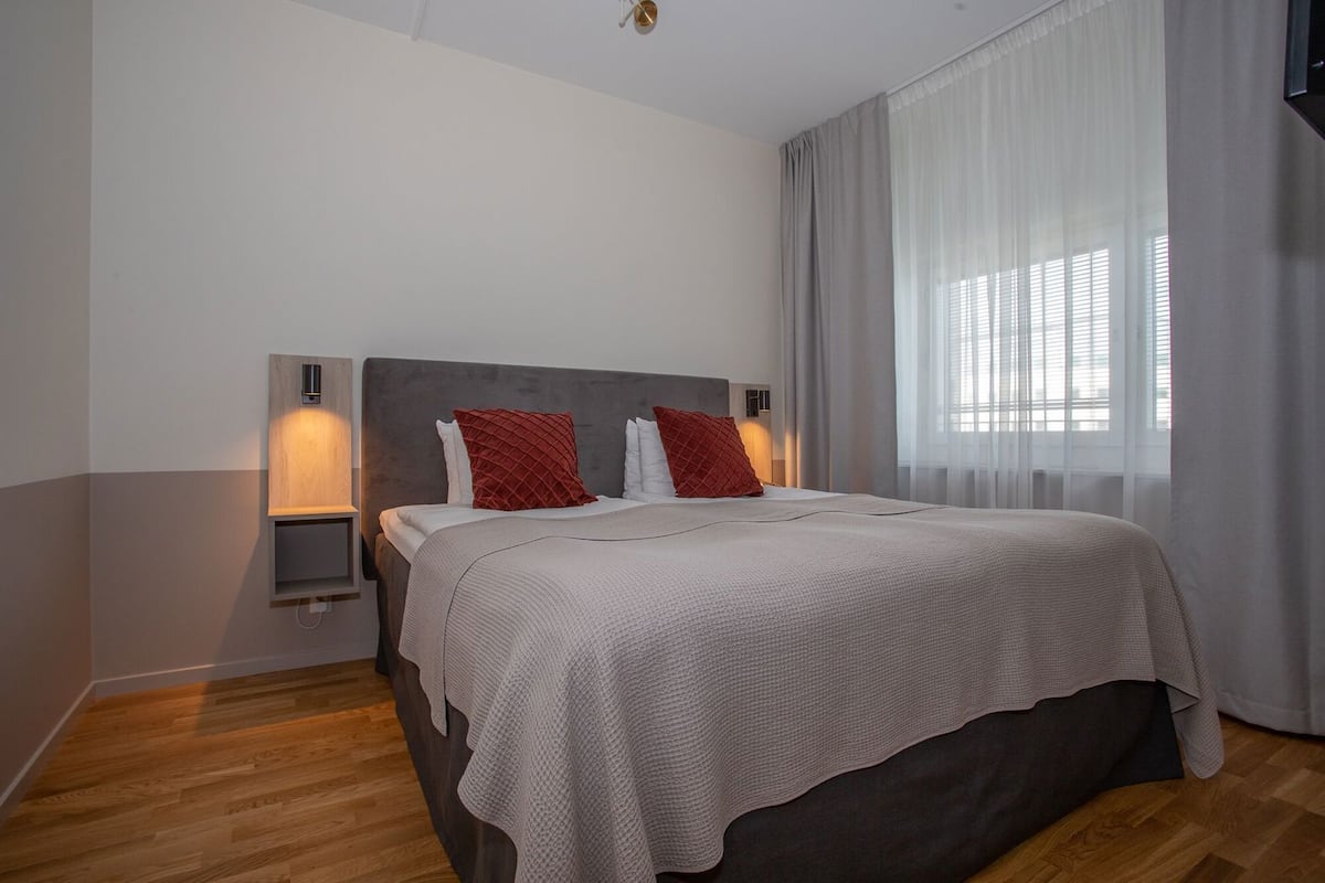 A bedroom with a bed, a bedside table, a wall-mounted light, and a window with curtains.