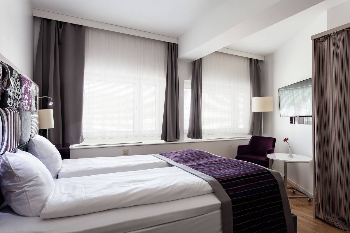 A hotel room with two beds, a small table, a chair, and a window with curtains.