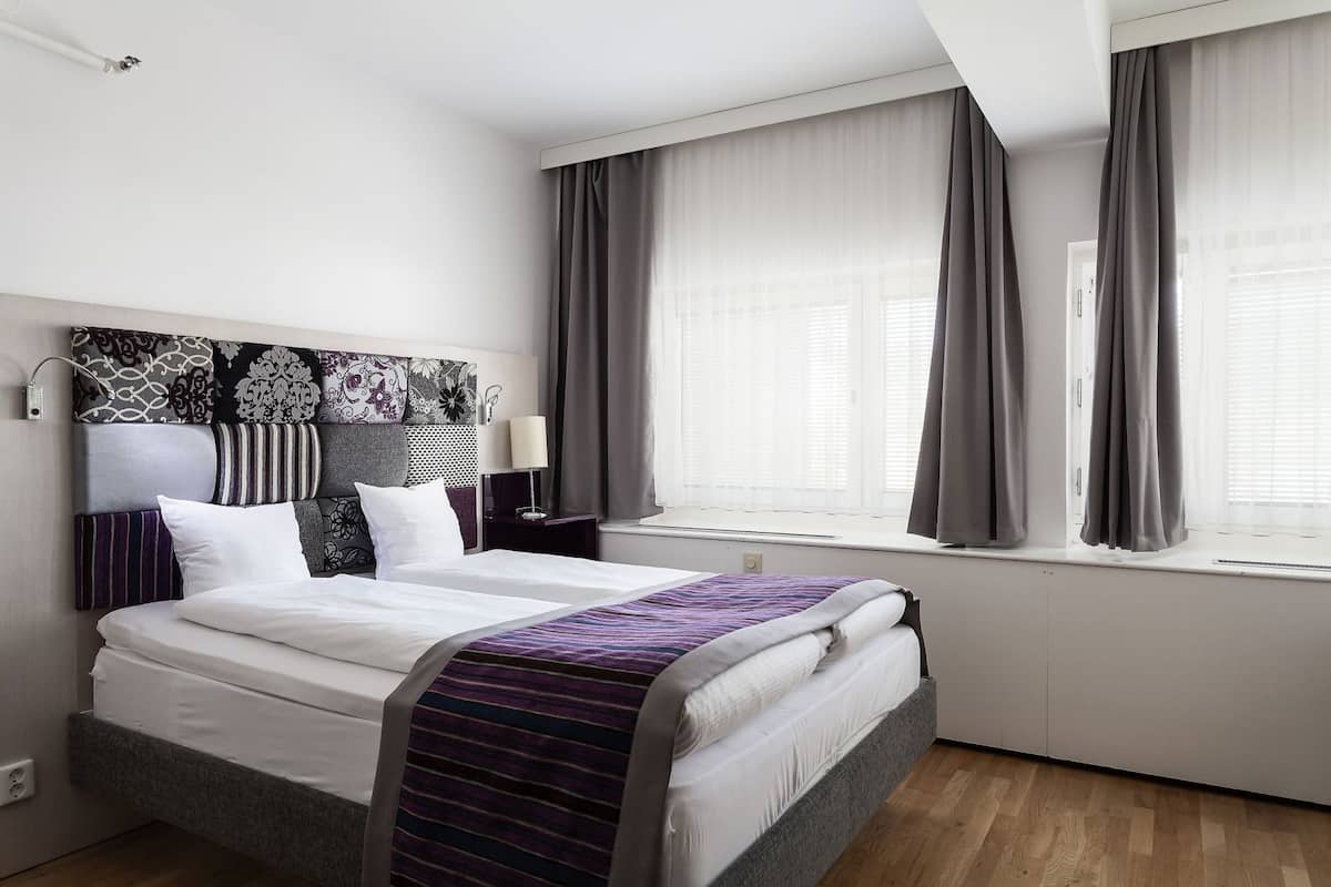 A modern hotel room with a neatly made bed, a headboard with patterned designs, a bedside table with a lamp, and large windows with curtains.