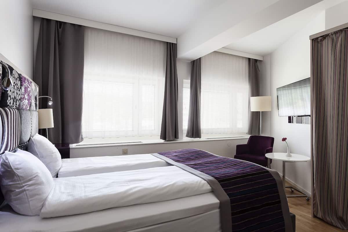 A hotel room with two beds, a small table, a chair, and a window with curtains.