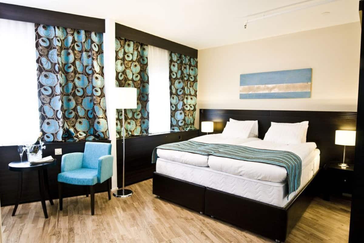 A hotel room with a bed, bedside tables, a chair, a floor lamp, and a wall with a decorative pattern.