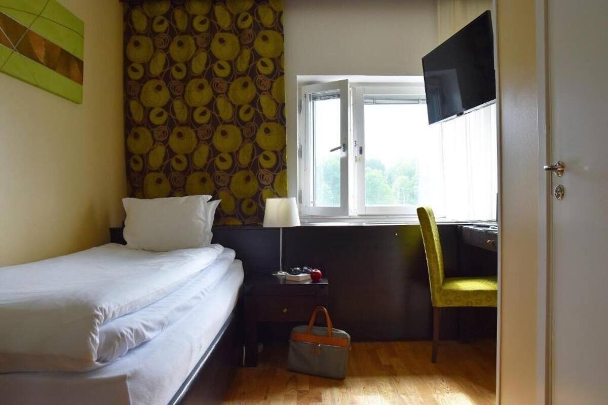 A hotel room with a bed, a desk with a chair, a window with a TV, and a patterned wall.
