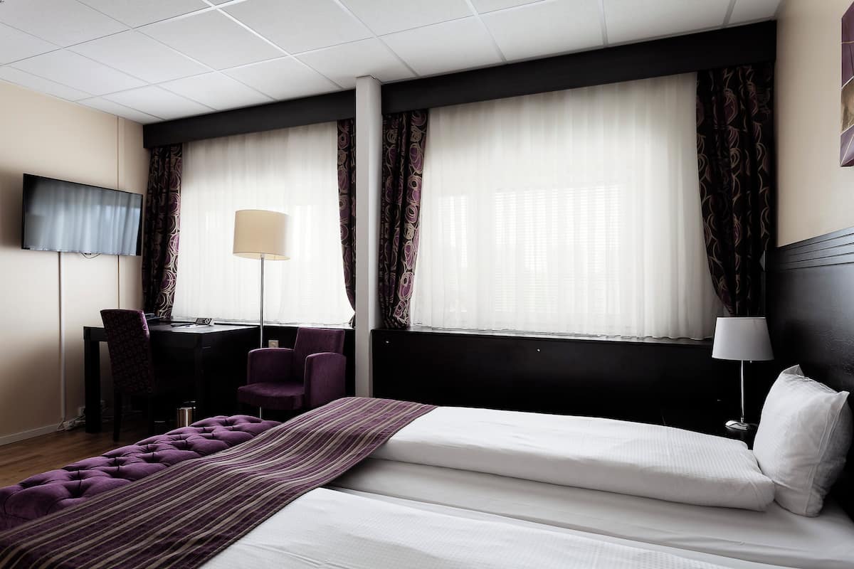 A hotel room with a bed, a desk, a chair, a TV, and a window with curtains.