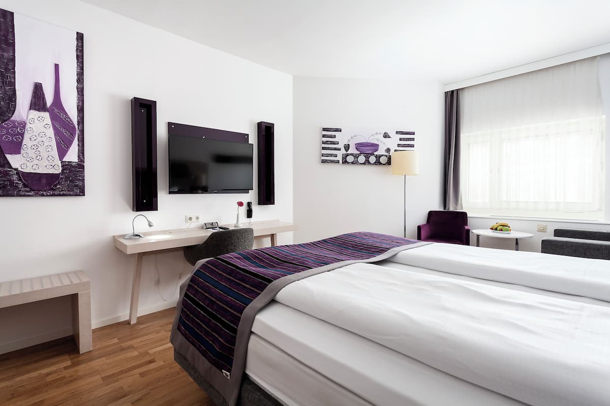 A hotel room with a bed, a desk, a chair, a TV, and artwork on the walls.