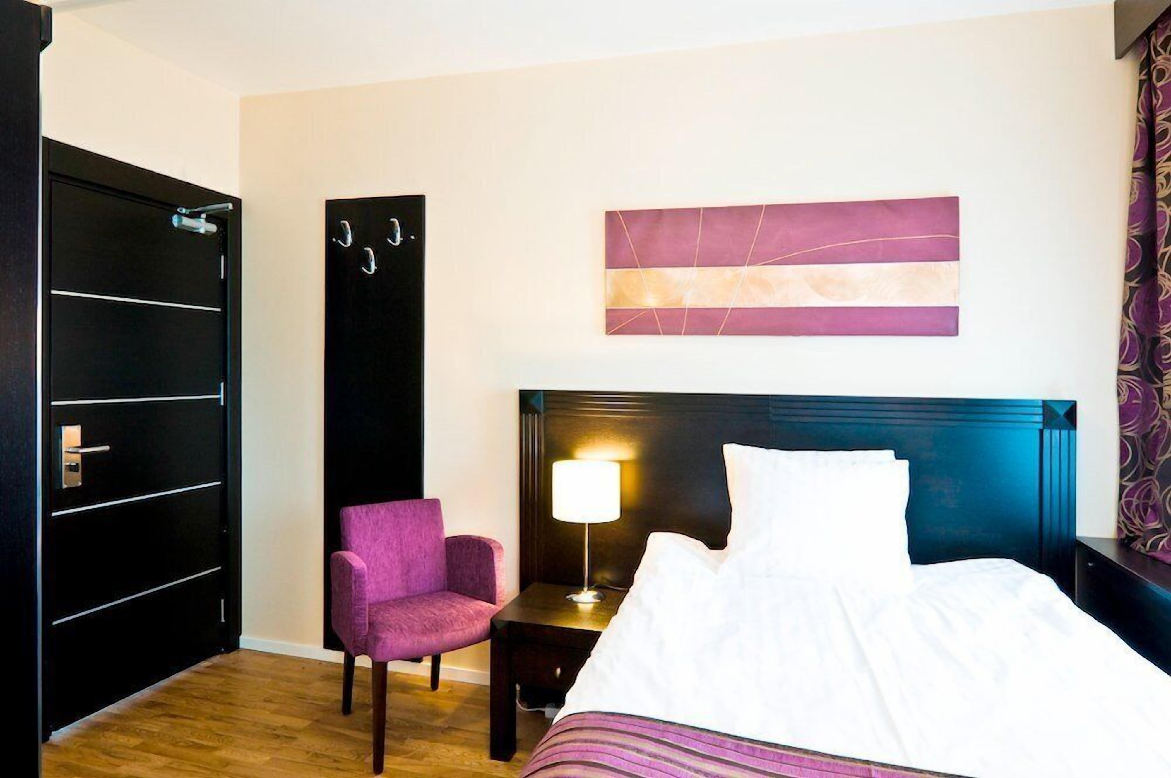 A hotel room with a bed, a nightstand with a lamp, a chair, a wardrobe, and a wall art piece.