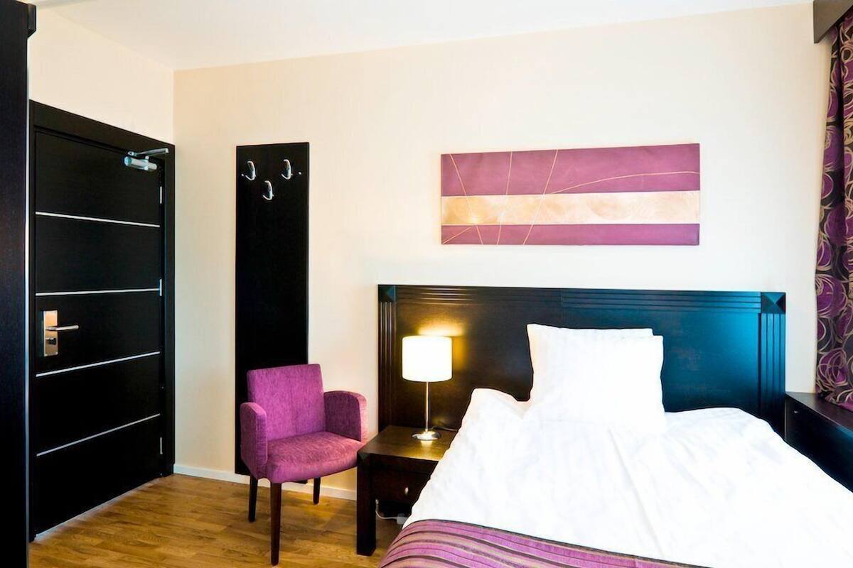 A hotel room with a bed, a nightstand with a lamp, a chair, a wardrobe, and a wall art piece.