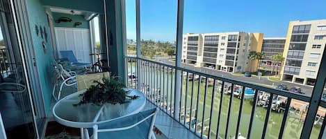 A balcony with a table, chairs, and a potted plant. It overlooks a marina with boats and buildings in the background.