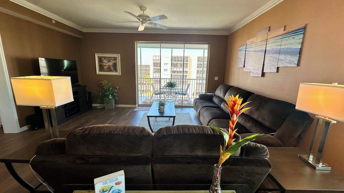 A living room with a sofa, a coffee table, a TV, and a balcony with a view of buildings.