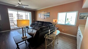 A living room with a black leather sofa, a glass coffee table, a ceiling fan, and a large window with a view of buildings. - Casa Marina 424 - Dockside Living (Fort Myers Beach)