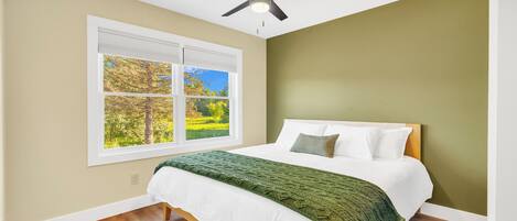 Un dormitorio con una cama, un ventilador de techo, una ventana con vistas a árboles y suelo de madera.