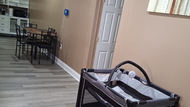 A room with a crib, a dining table with chairs, and a kitchen area in the background.