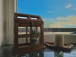 Um candeeiro de madeira com uma planta no interior, uma vela e um tabuleiro sobre uma superfície de mármore perto de uma janela.